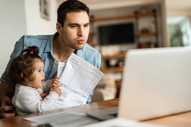 giovane padre che legge la posta su laptop mentre fa da babysitter a casa. - pensive only baby girls baby girls baby foto e immagini stock