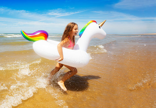 Happy smiling girl run over waves in the swimming inflatable unicorn on the sand sea beach