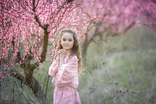 A girl holds her heart-shaped hands against a tree background. A child in a garden with flowering peach trees