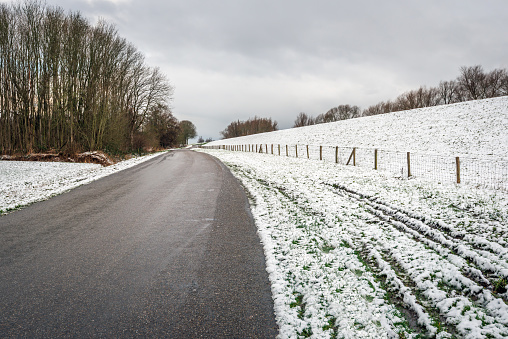 Seemingly endless asphalt road at the bottom of a snow-covered Dutch dike with a long fence made of wooden posts with mesh.