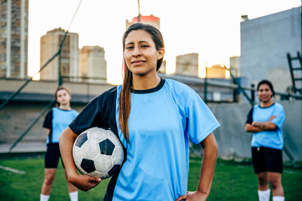 Confident young Hispanic footballer identifying as non-binary Close-up of Buenos Aires player in late 20s with ball under arm and football teammates in background. gender fluid photos stock pictures, royalty-free photos & images