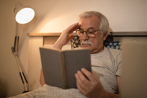 Pensive mature man resting in bed and reading a book at night.