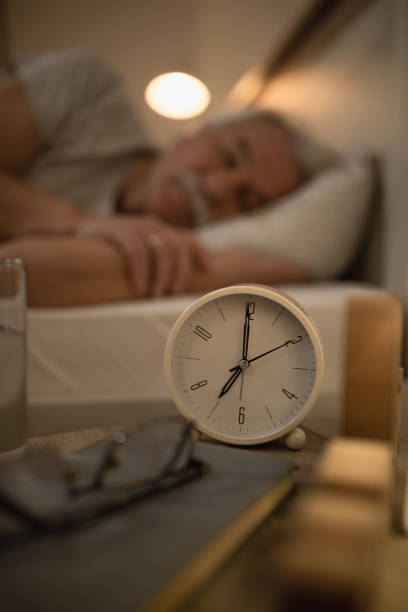 istock Alarm clock on bedside table with man sleeping in the background. 1210141011