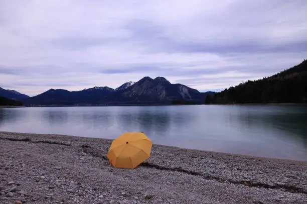 A yellow umbrella at the lakeside in a mountain range. Gloomy mood, grey cloudy sky. Mountain silhouette at the horizon. Pebbles and a bright umbrella in the foreground. No persons. Tranquil scene.