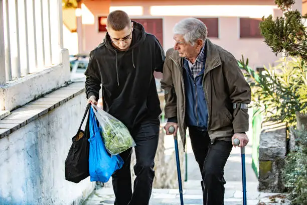 Young Adult Man Helping Senior Adult Man With Crutches To Walk and Carry His Groceries.