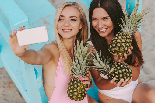Happy young girls in swimsuits making sunny photos on smart phone camera at seaside. Beautiful cheerful women friends posing with pineapples in hands and taking selfie together on summer beach