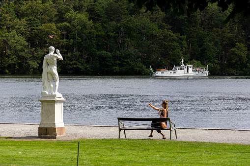 Stockholm, Sweden - July 13, 2019: A woman sitting on a bench next to a statue and water taking a selfie in the wonderful surroundings called Drottningholm