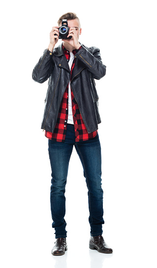 Front view of aged 18-19 years old with brown hair caucasian boys tourist standing in front of white background wearing plaid shirt who is laughing who is photographing and holding camera