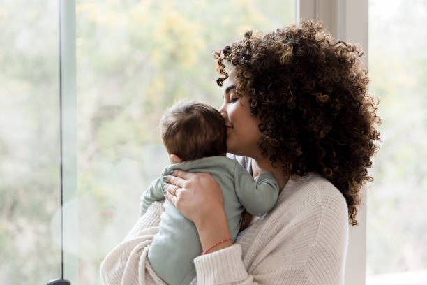 Mom spends time with baby boy Young mom gives her baby a kiss on his head while standing next to a window in their home biracial newborn stock pictures, royalty-free photos & images