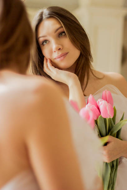 Closeup of young beautiful woman looking at her reflection in mirror and holding bouquet of spring flowers. Pretty and tender girl in white dress posing against mirror with tulips in hands. 8 march