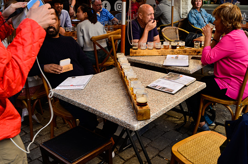 Bruges, Belgium. August 2019. Tasting of Belgian craft beers: twelve mugs arranged on the wooden cup holder. People are sitting at the tables enjoying. A boy photographs them with his smartphone.