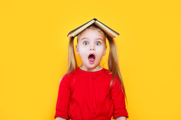 Shocked little girl holding book on her head and looking at camera stock photo