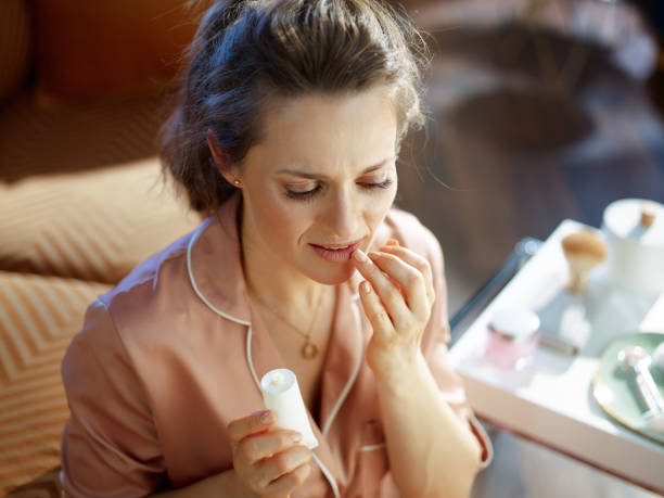 woman with white cosmetic tube applying lip contour cream stylish woman in pajamas with white cosmetic tube applying lip contour cream near table with toiletries in the modern house in sunny winter day. herpes stock pictures, royalty-free photos & images