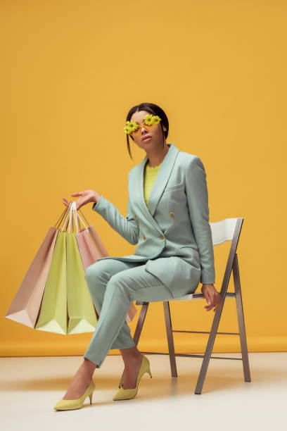 привлекательная афро-американская женщина в костюме и солнцезащитных очках с цветами, держащими сумки для покупок, сидя на желтом - sitting flower female studio shot стоковые фото и изображения