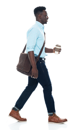 Profile view of aged 20-29 years old with curly hair african ethnicity male walking in front of white background wearing satchel - bag who is laughing and holding coffee cup