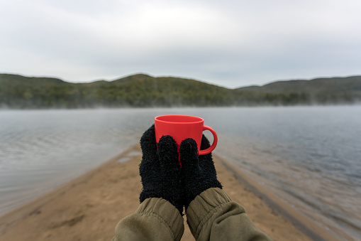 A young women is drinking a coffee by the lake in autumn in camping. She is contemplating the view and you can see from her personal perspective. She is wearing warm clothing.