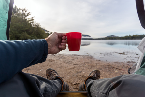 A man is drinking a coffee by the lake in camping. He is sitting in his tent and contemplating the view. You can see his personal perspective of the nature surrounding him.