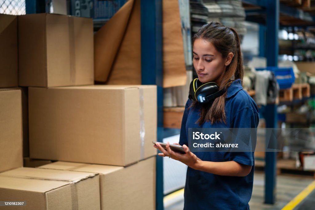 Warehouse worker updating the stock on mobile app Female warehouse worker updating the stock on mobile phone app. Woman in uniform working in a factory warehouse. Warehouse Stock Photo