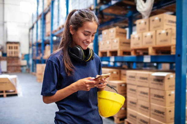 Woman warehouse worker using mobile phone Young woman using her mobile phone in large warehouse. Female warehouse worker reading text message on her cell phone. blue collar worker stock pictures, royalty-free photos & images
