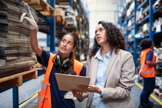 Manager with foremen controlling stock in warehouse. Warehouse worker pointing at the rack with a businesswoman holding a clipboard.