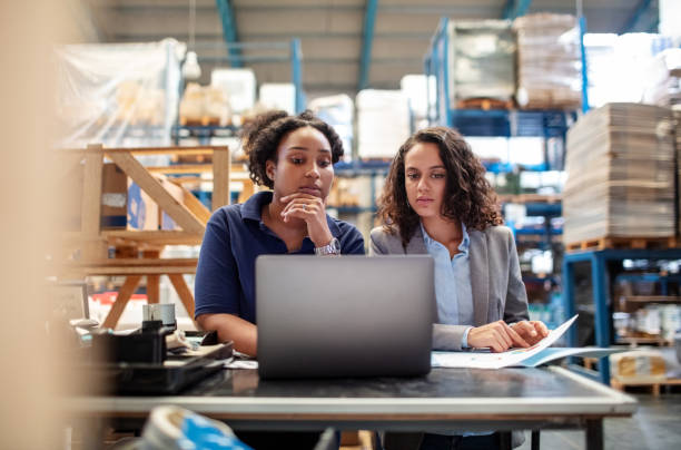 female worker with supervisor working on laptop - freight transportation warehouse manufacturing shipping imagens e fotografias de stock