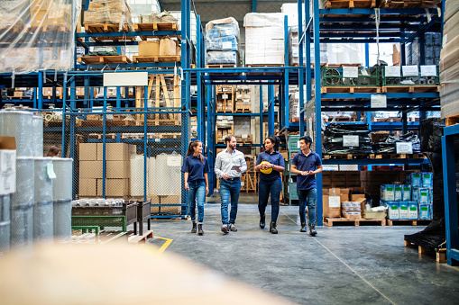 Group of four warehouse employees walking by storage racks. Warehouse employees walking through aisle and talking.