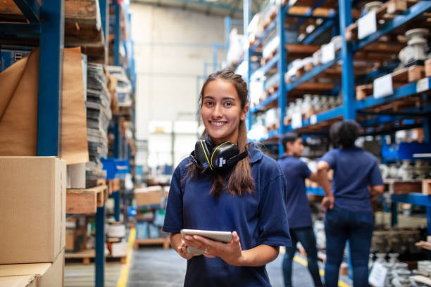 Confident female warehouse worker Portrait of a female factory employee with a digital tablet looking at camera and smiling. Woman warehouse worker with people in background. hardware store photos stock pictures, royalty-free photos & images