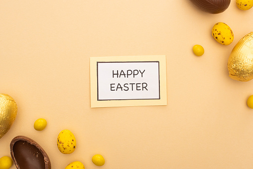 Top view of card with happy easter lettering, chocolate eggs, candies and painted quail eggs on beige background