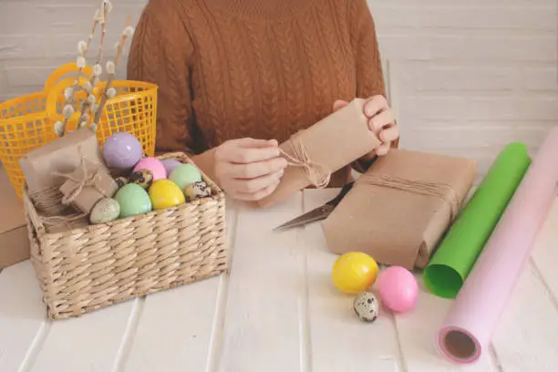 Photo of Young woman wrapping easter gifts. Colored eggs and holiday symbols on a light table. Zero waste concept.