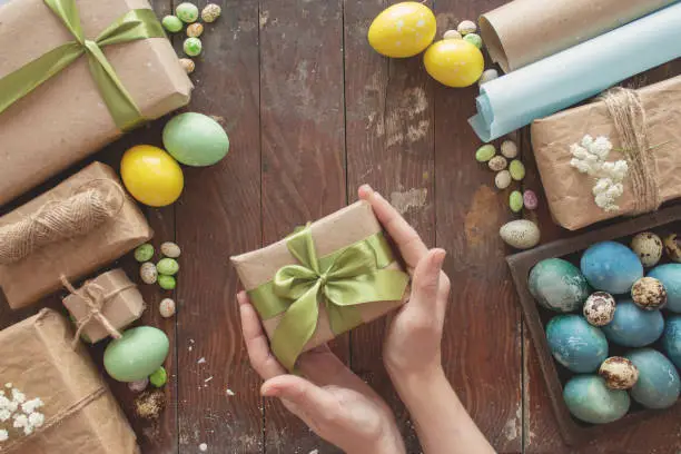 Photo of A young woman holds a gift in her hands. Colored eggs and holiday symbols on a dark wooden table. Zero waste concept.