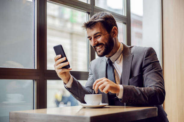 junge lächelnde bärtige geschäftsmann sitzt in einem café, lesen etwas lustiges auf dem smartphone und rühren kaffee. - rühren stock-fotos und bilder