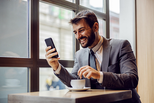 Joven hombre de negocios barbudo sonriente sentado en la cafetería, leyendo algo divertido en el teléfono inteligente y agitando el café. photo