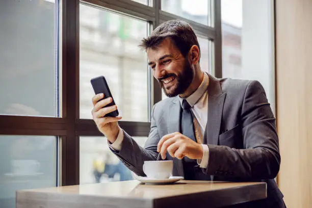 Photo of Young smiling bearded businessman sitting in coffee shop, reading something funny on smart phone and stirring coffee.