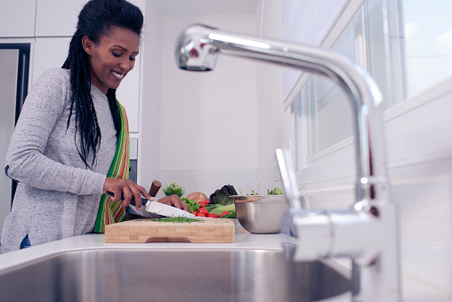 Happy young woman chopping vegetables for a healthy meal. The woman standing in her new modern kitchen and preparing salad on the countertop. Sink and faucet are on the foreground.