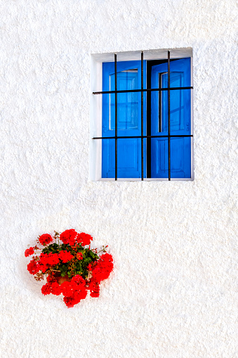 A blue wooden shuttered window of a white washed spanish town house, with red potted plants against the white washed wall.