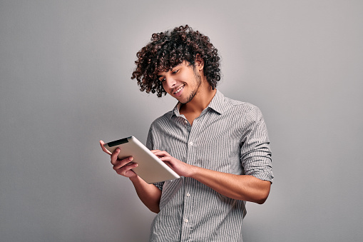 Smiling arabian young man looking at his digital tablet screen, standing against gray wall. Isolated image