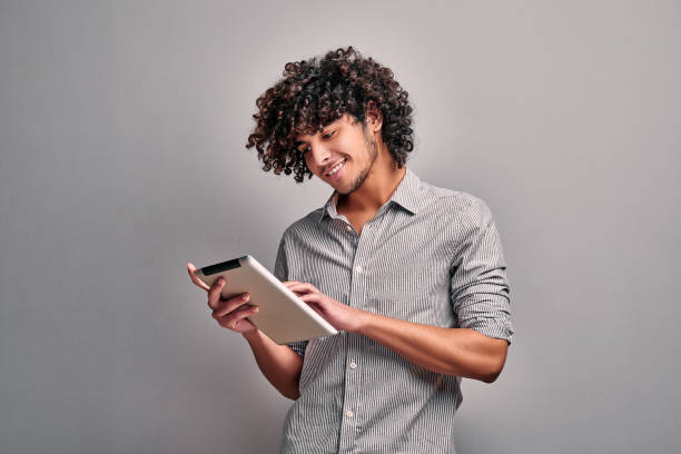Smiling arabian young man looking at his digital tablet screen Smiling arabian young man looking at his digital tablet screen, standing against gray wall. Isolated image tablet stock pictures, royalty-free photos & images