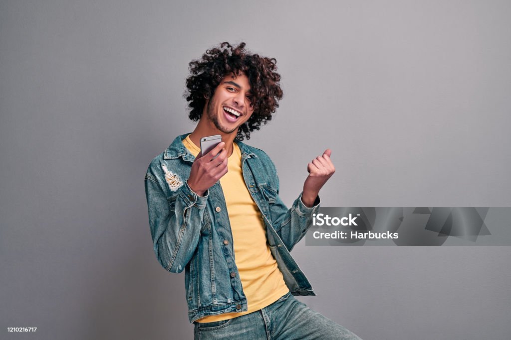 Joyful student with phone Handsome eastern excited man dancing with smartphone in hand on isolated gray background. Joyful student with phone Men Stock Photo