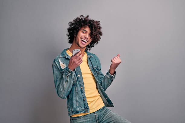 Joyful student with phone Handsome eastern excited man dancing with smartphone in hand on isolated gray background. Joyful student with phone jeans photos stock pictures, royalty-free photos & images