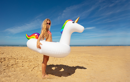 Happy little blond girl stand with inflatable unicorn on the sand beach