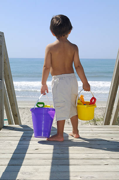 Child heading to the beach with toys Child heading to the beach with toys.  A little boy walking down a beach house boardwalk to the beach.  He is carrying a yellow bucket and a purple bucket.  Both buckets have beach toys in them. emerald isle north carolina stock pictures, royalty-free photos & images
