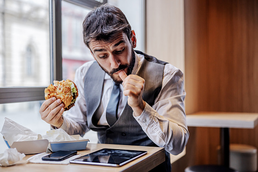 Hungry man in suit sitting in fast food restaurant on lunch break, eating cheese burger and reading news on tablet.