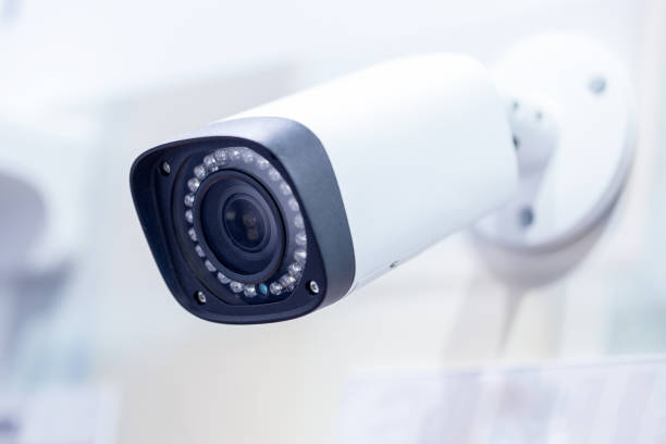istock Big white professional surveillance camera. CCTV mounted on ceiling. LED IR lights around lens. Security system concept. Copyspace, neutral light blue backgound 1210266874