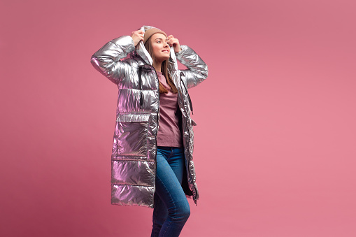 fashionable and modern young woman in a puffy light down jacket throws a hood over her head. The jacket is a silver metallic color. Girl in street clothes, jeans and sneakers, on a pink background