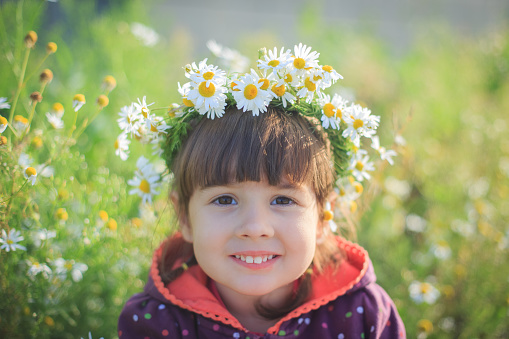 A little girl in a wreath of daisies smiles and sits in the green grass in summer. Sunny day.