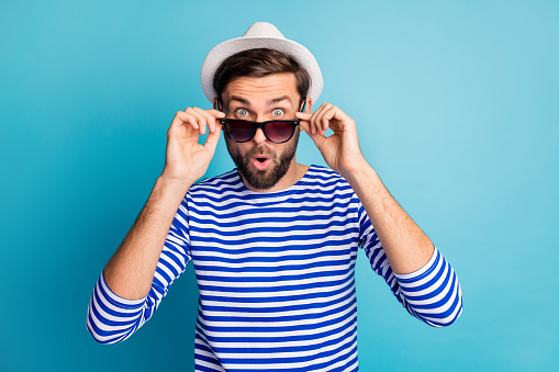 Photo of funky excited handsome guy tourist taking off cool black sun specs, open mouth read discounts banner wear striped sailor shirt cap isolated blue color background