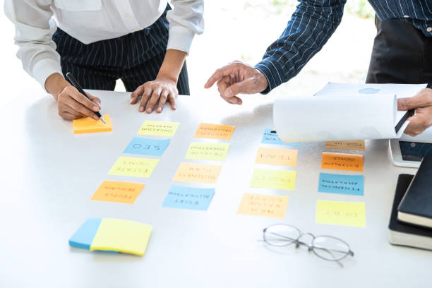 istock Two creative business people meeting and planning use post it notes sticky note on desk to share idea, Analysis data chart and graph with teamwork strategy brainstorming in office 1210303521