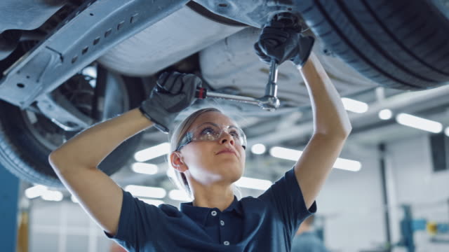 Slow Motion Portrait Footage of a Female Mechanic Working on a Vehicle in a Car Service. Empowering Woman Wearing Gloves and Using a Ratchet Underneath the Car. Modern Clean Workshop.