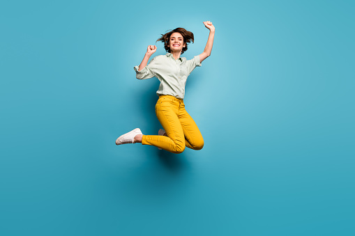 Full body photo of funky pretty lady jumping high up celebrating weekend vacation start wear casual green shirt yellow pants sneakers isolated blue color background