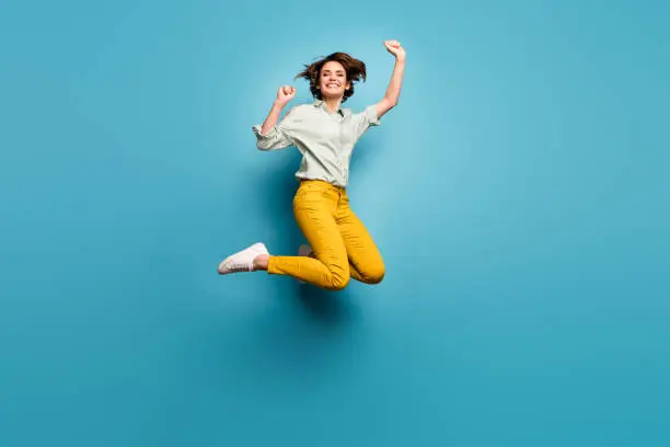 Full body photo of funky pretty lady jumping high up celebrating, weekend vacation start wear casual green shirt yellow pants sneakers isolated blue color background
